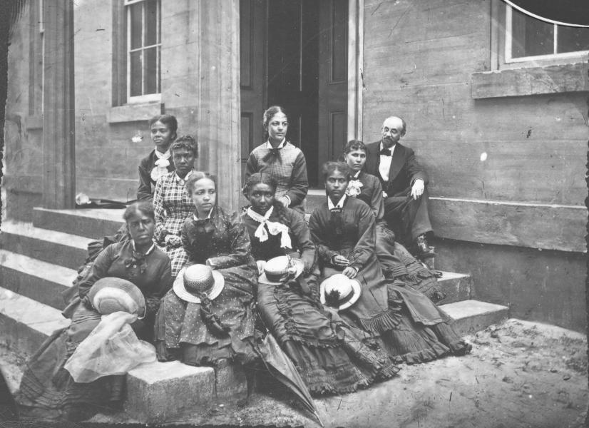 Eight well-dressed Black women and one Black man sit on steps, looking serious.