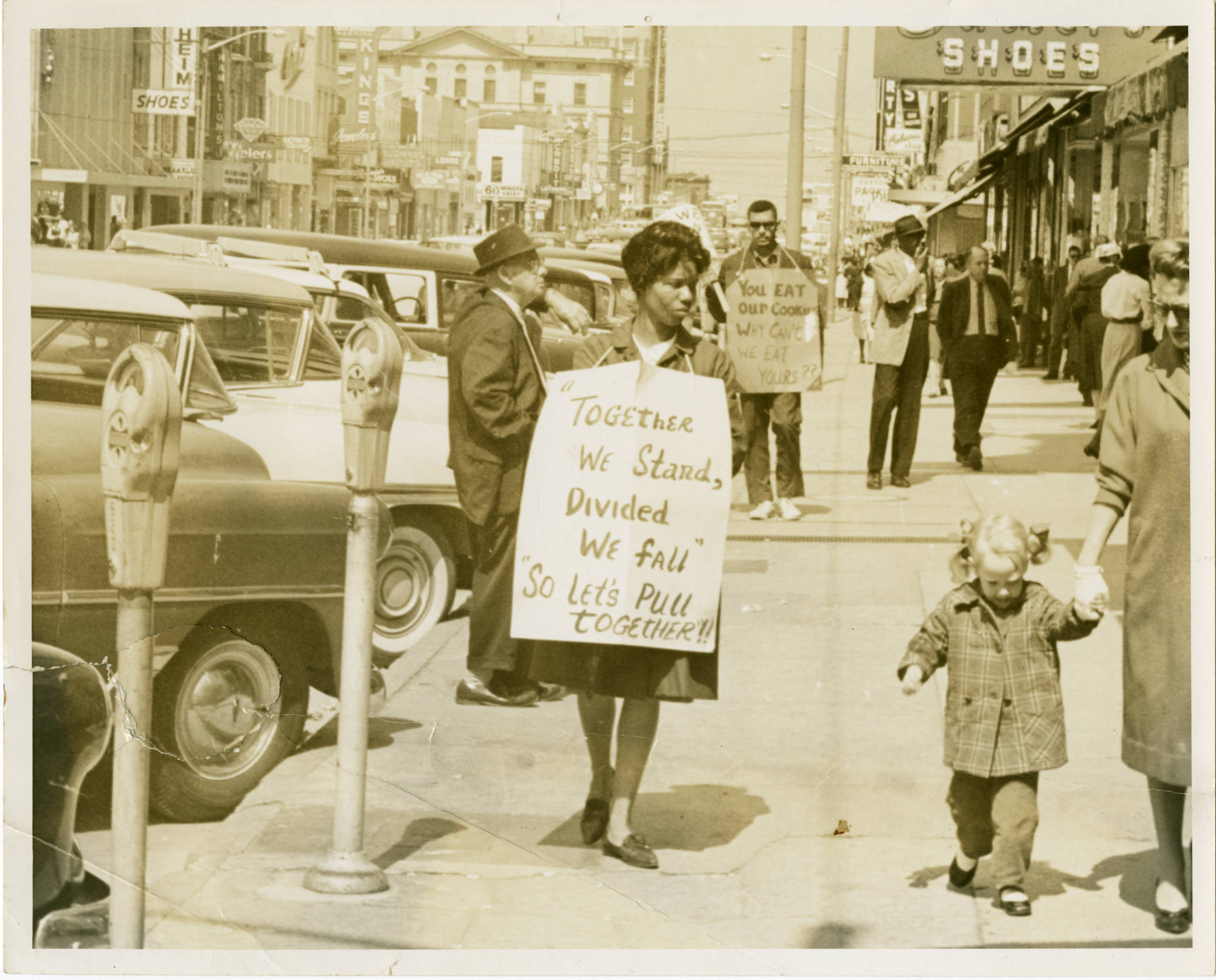 Two protesters carrying signs walk down a busy Main Street sidewalk.