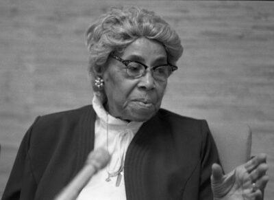 An older African American woman with short gray hair, glasses, and a nice jacket speaks into a microphone.
