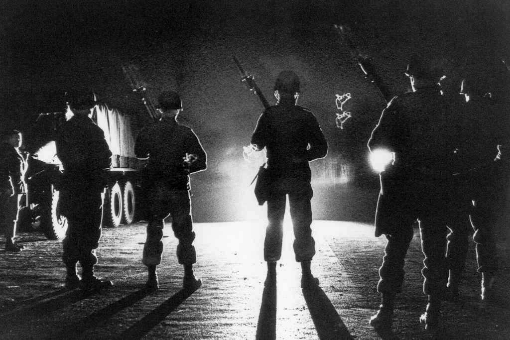 Silhouettes of armed National Guardsmen lit by headlights.